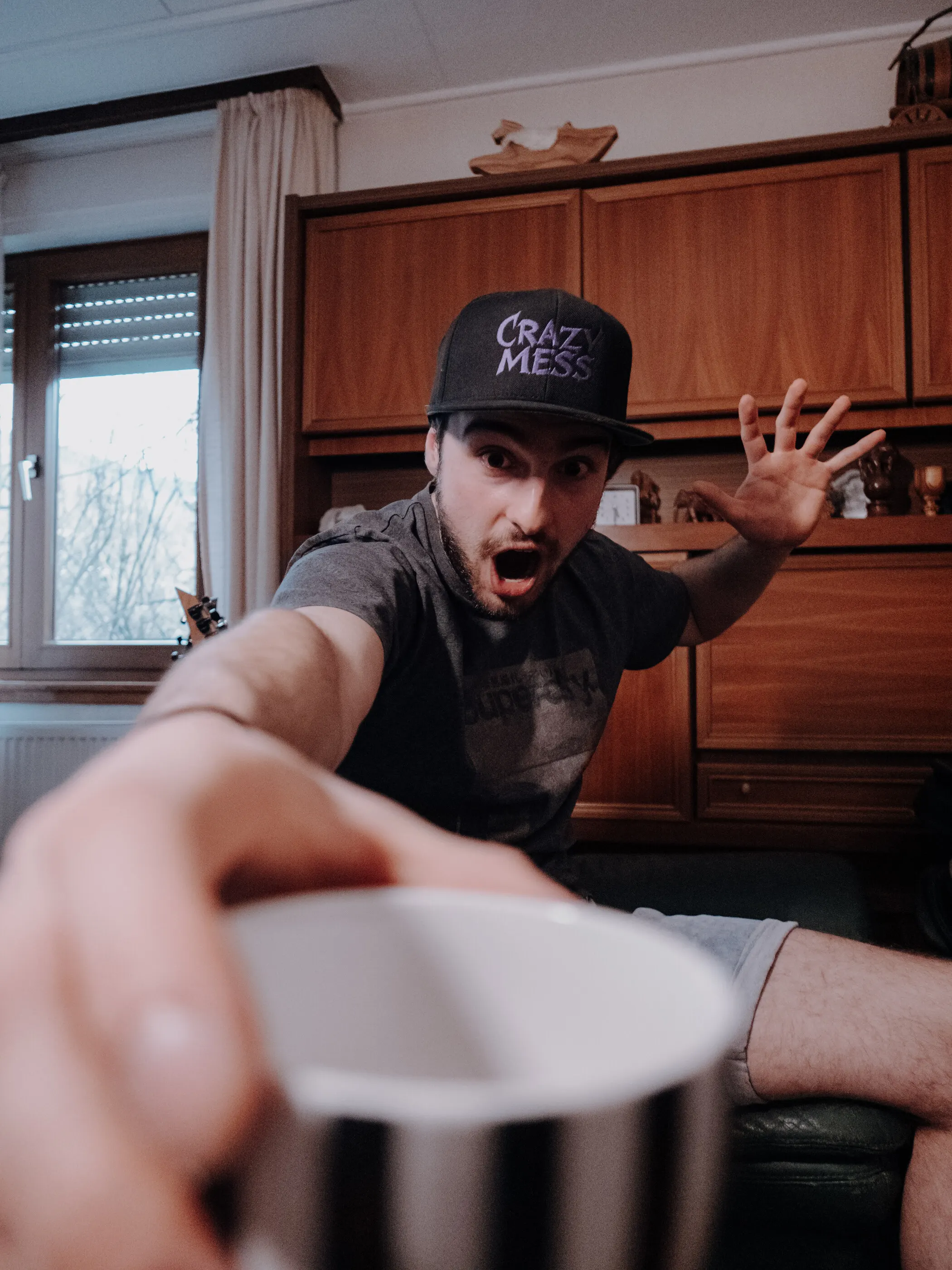 Mann mit Cap streckt der Kamera lachend eine Kaffeetasse entgegen
